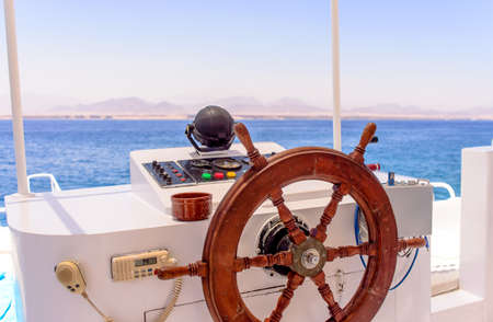 Ships wooden spooked wheel and navigation console at the helm of a yacht or motorboat cruising mid ocean with a distant coastline visible on the horizon, nobody in viewの写真素材