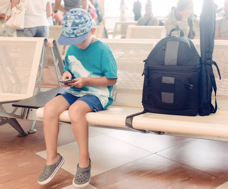 Little boy sitting in an airport departure hall contentedly playing on his tablet or mobile phone as he waits for his flight with his luggageの写真素材