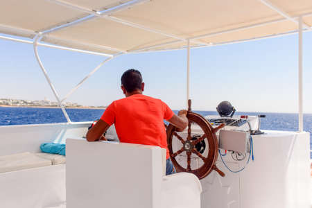 Captain or helmsman on a yacht or motorboat cruising mid ocean viewed from behind sitting at the helm in a bright red shirtの写真素材