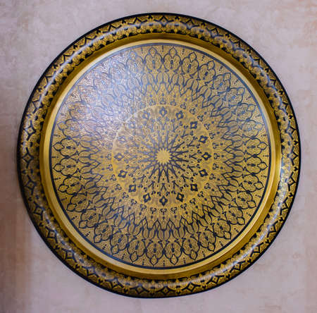 Ornate engraved round brass wall plaque mounted on a white wall with a geometric repeat pattern,...