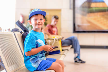 Little boy sitting in an airport departure hall contentedly playing on his tablet or mobile phone as he waits for his flight with his luggageの写真素材