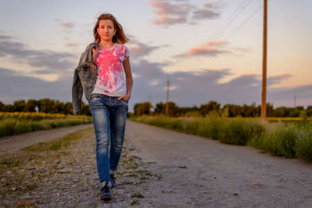 Young girl taking an evening walk in the country strolling down a rural road bordered with fields of sunflowers towards the cameraの写真素材