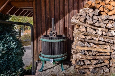 Old fashioned wine press next to pile of cut logs next to wall near garage with tree and gutterの写真素材