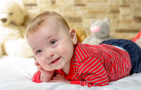 Cute chubby little baby with a happy smile lying on its stomach on a bed with a row of soft cuddly...