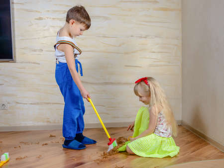 Cute little boy and girl cleaning up in the house using a toy plastic broom and pan to sweep up wood shavings on a parquet floor looking at the camera with happy smilesの写真素材
