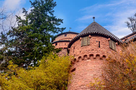 Beautiful red brick castle of Haut-Koenigsbourg in Alsace, France with conical spires and closed windows, surrounded by foliage under blue sky and white cloudsのeditorial素材