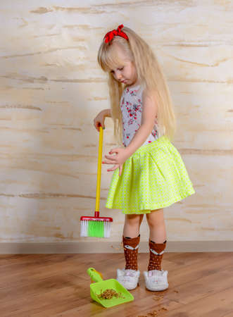 Pretty little blond girl cleaning the house with a colorful plastic toy broom and pan bending forward as she carefully sweeps up the dirt, full length close upの写真素材