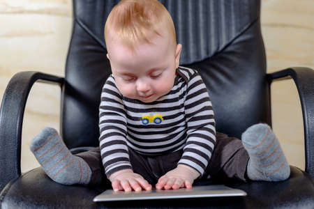 Cute Little Boy Playing with his Tablet Computer While Sitting on a Black Office Chairの写真素材