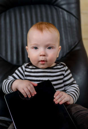 Adorable Baby Boy Holding his Tablet Computer While Sitting on an Office Chair and Looking at the...