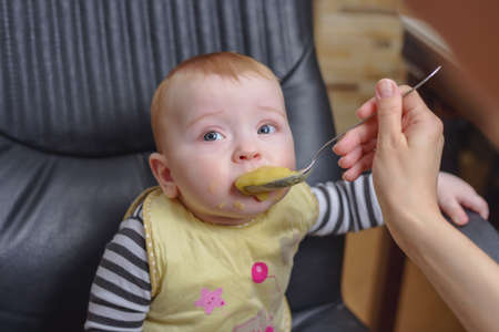 Cute Baby Boy Sitting on a Black Office Office Chair, Eating Porridge While Looking Away from the Camera.の写真素材