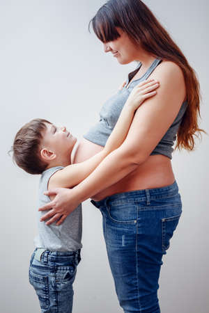Side view of grinning little boy in blue jeans hugging pregnant mom with long hair over gray backgroundの写真素材