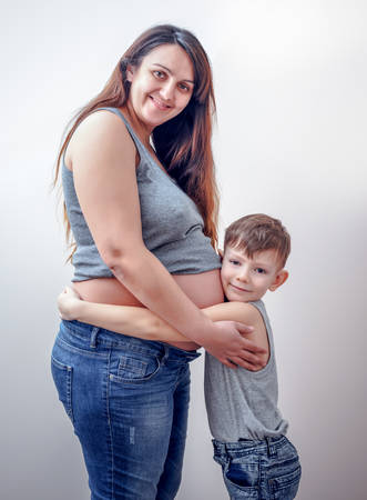 Brown haired expectant young woman with her belly exposed in blue jeans wearing red headband embracing her young childの写真素材