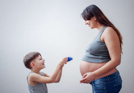 Cute happy boy giving smiling pregnant woman with large belly and long hair a gift over gray background with copy spaceの写真素材