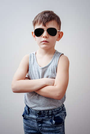 Cute little single boy in sunglasses, sleeveless shirt and blue jeans with folded arms over gray backgroundの写真素材