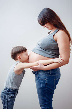 Side view of cute little boy in blue jeans kissing belly of obscured pregnant mom with hands on hipsの写真素材