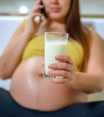 Healthy and relaxed woman in the third trimester of pregnancy sitting on a couch at home while talking on mobile phone and drinking a glass of milkの写真素材