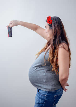 Side view of single pregnant lady in long hair taking a self portrait while holding camera phone in front of herselfの写真素材