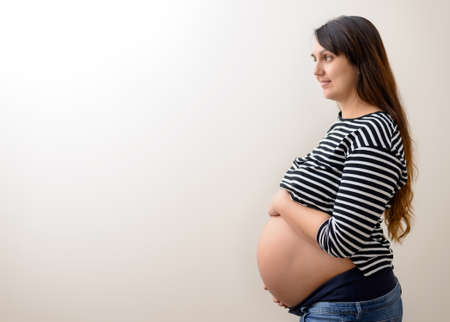 Calm pregnant young woman with long hair in black and white striped shirt holding belly and grinning...
