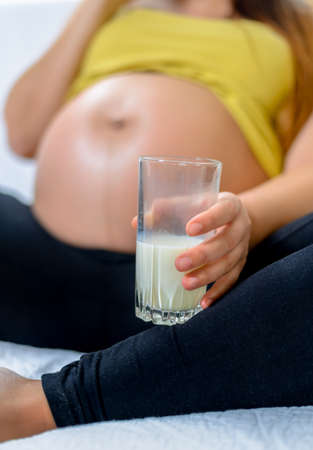 Pregnant woman holding a healthy glass of farm fresh milk rich in calcium, close up of her bare belly and handの写真素材