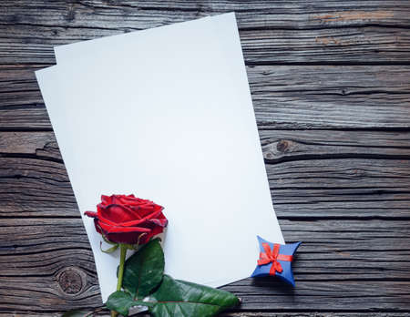 Pair of stacked blank papers centered on table with red rose and little blue gift boxの写真素材