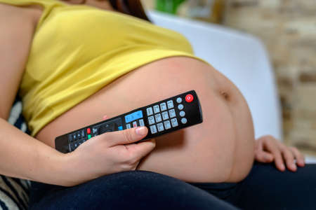 Pregnant woman relaxing watching television at home on a sofa changing channels with the remote...