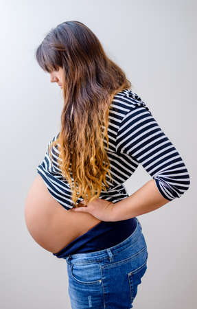 Heavily pregnant woman standing sideways looking down at her bare belly, isolated on white
