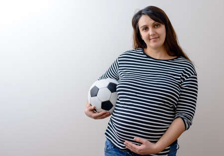 Grinning pregnant woman in striped shirt with football ball looking like a soccer momの写真素材