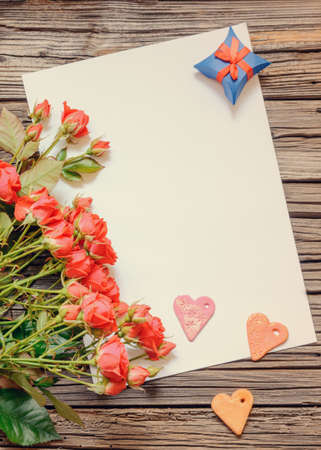 Single white blank sheet of paper with copy space on table with weathered wooden surface next to bundle of pink roses, heart shapes and little gift boxの写真素材
