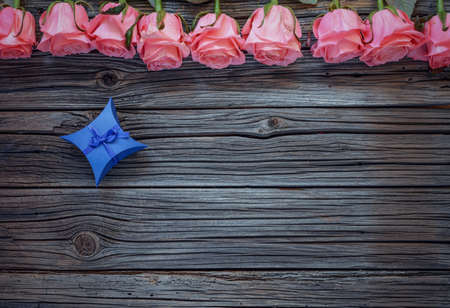 Tiny blue diamond shaped bowed gift box over worn out wooden background with roses on topの写真素材