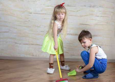 Cute little boy in blue pants, slippers and suspenders using toy broom and dustpan with girl in green skirt on hardwood floorの写真素材