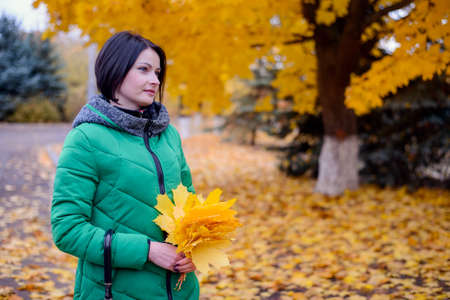 Cute young smiling woman in green winter coat holding large yellow maple tree leaves and umbrella outdoors in autumn sceneの写真素材