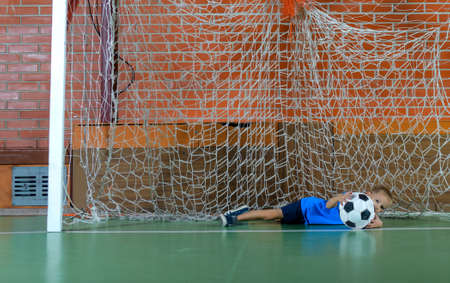 Young goalie saving a goal on an indoor soccer court lying across the goals on his side clutching the ballの写真素材