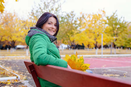 Cute young grinning woman in green winter coat sitting on bench with hand under chin near yellow trees in park during autumnの写真素材