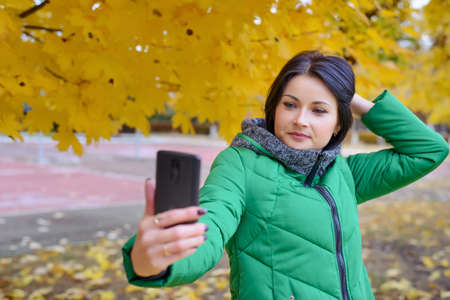 Gorgeous young woman in thick winter coat taking picture of herself at park with camera phone under...