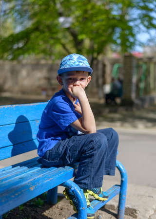 Patient young boy sitting waiting on a blue wooden outdoor bench in a trendy blue outfit with his hand to his chin and a bored expressionの写真素材