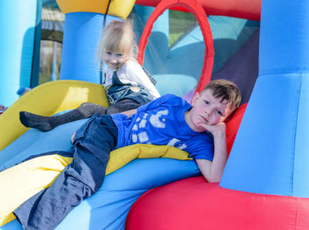 Cute young brother and sister having fun as they lay down on inflatable blue, yellow and red castle and slideの写真素材