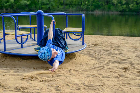 Little boy on a spinning merry-go-round waving at the camera as he looks back over his shoulder outdoors on a sandy beach near a watercourseの写真素材
