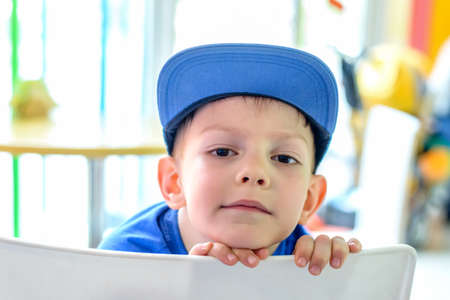 Handsome small boy in a blue baseball cap leaning his chin on the back of the chair giving the...