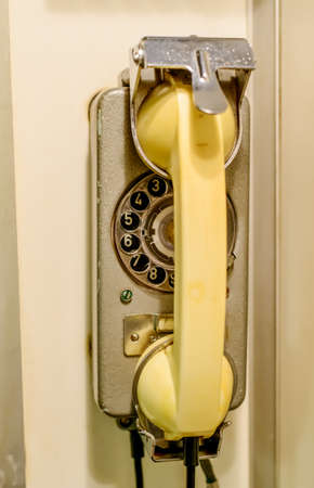 Old yellow wall-mounted rotary dial-up telephone in a cradle, close up view in a telecommunications...