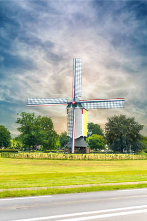 Colorful wooden windmill with four sails in an agricultural field alongside a highway against a cloudy blue skyの写真素材