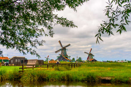 Leaves from trees surrounding view of old windmills with large bases at farm under partly cloudy skiesの写真素材