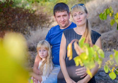 Young family enjoying a day in the mountains with a loving father hugging his pregnant wife and little daughter between tree foliageの写真素材