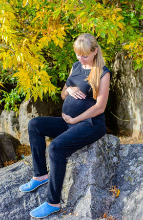 Heavily pregnant woman with a lovely smile sitting relaxing outdoors in the autumn sunshine tenderly holding her swollen bellyの写真素材