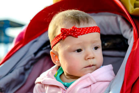 Contented happy little baby girl wearing a fun red headband and bow sitting in a push-chair laughing at the camera outdoors near a mountain lakeの写真素材