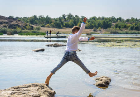 Small boy playing on rocks at the lake jumping from one to another with his arms outstretched and a smile of pleasureの写真素材