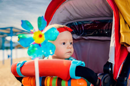 Cute little baby girl wearing a red headband sitting in a colorful push-chair or pram staring curiously at the cameraの写真素材