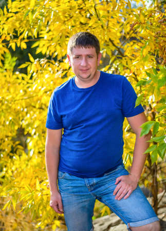 Friendly chubby man standing with his hands in his pockets in front of colorful yellow autumn foliage looking at the camera with a quiet smileの写真素材
