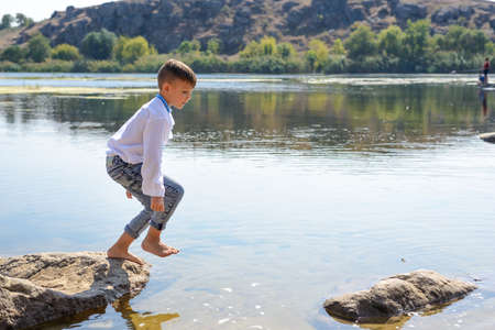 Small boy playing on rocks at the lake jumping from one to another with his arms outstretched and a smile of pleasureの写真素材