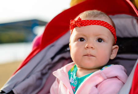 Curious cute little baby girl staring at the camera with an intense expression as she sits quietly in her push chair outdoors at a lakeの写真素材