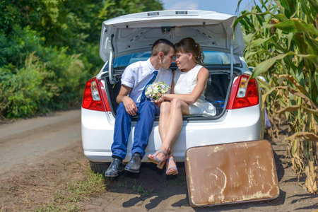 Unidentifiable married couple obscured by car trunk outside beside cornfield. Large brown suitcase on ground.の写真素材